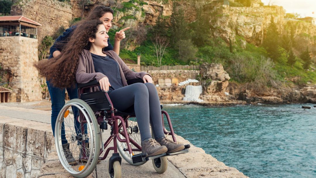 zwei Frauen, eine sitzt im Rollstuhl die stehende zeigt mit dem Finger in die Ferne
